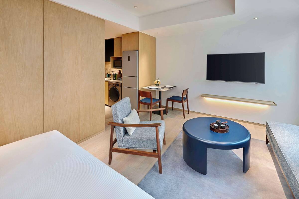 Une chambre d’hôtel moderne avec un grand lit, un coin salon comprenant deux fauteuils, une petite table ronde, un téléviseur à écran plat fixé au mur et, au fond, une kitchenette.