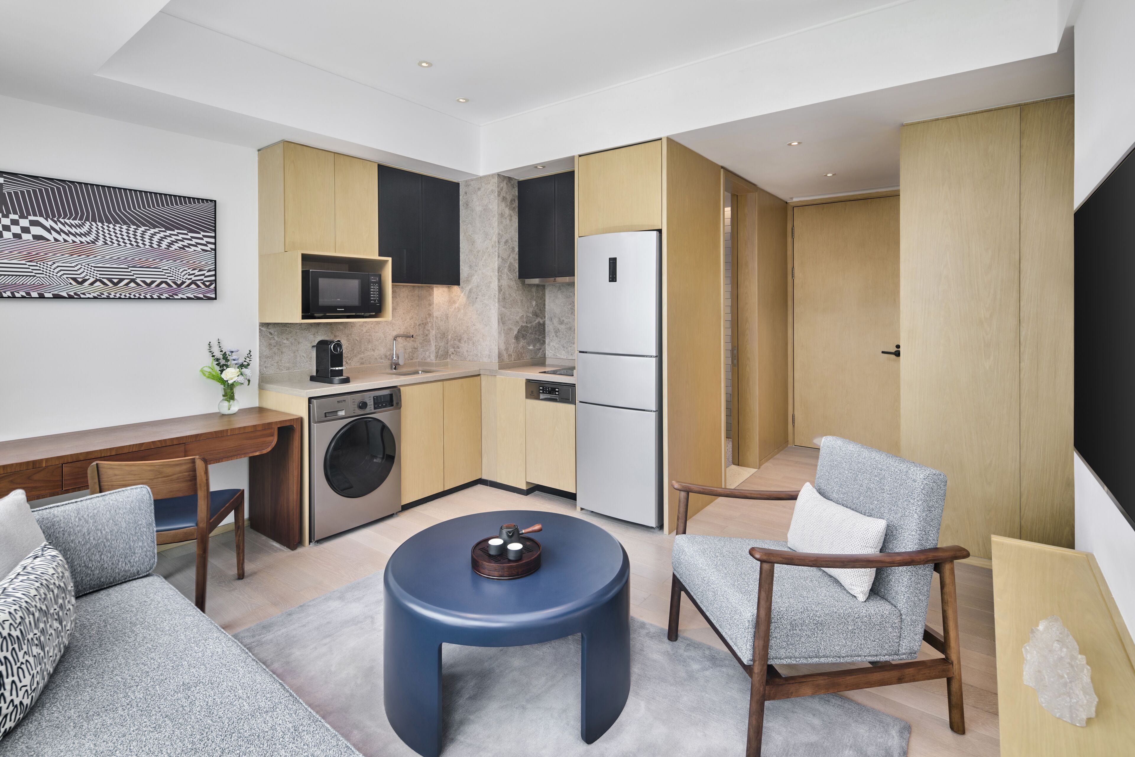 Una cocina moderna con microondas, horno y refrigerador. Hay una mesa redonda azul con una tetera y dos sillones grises.