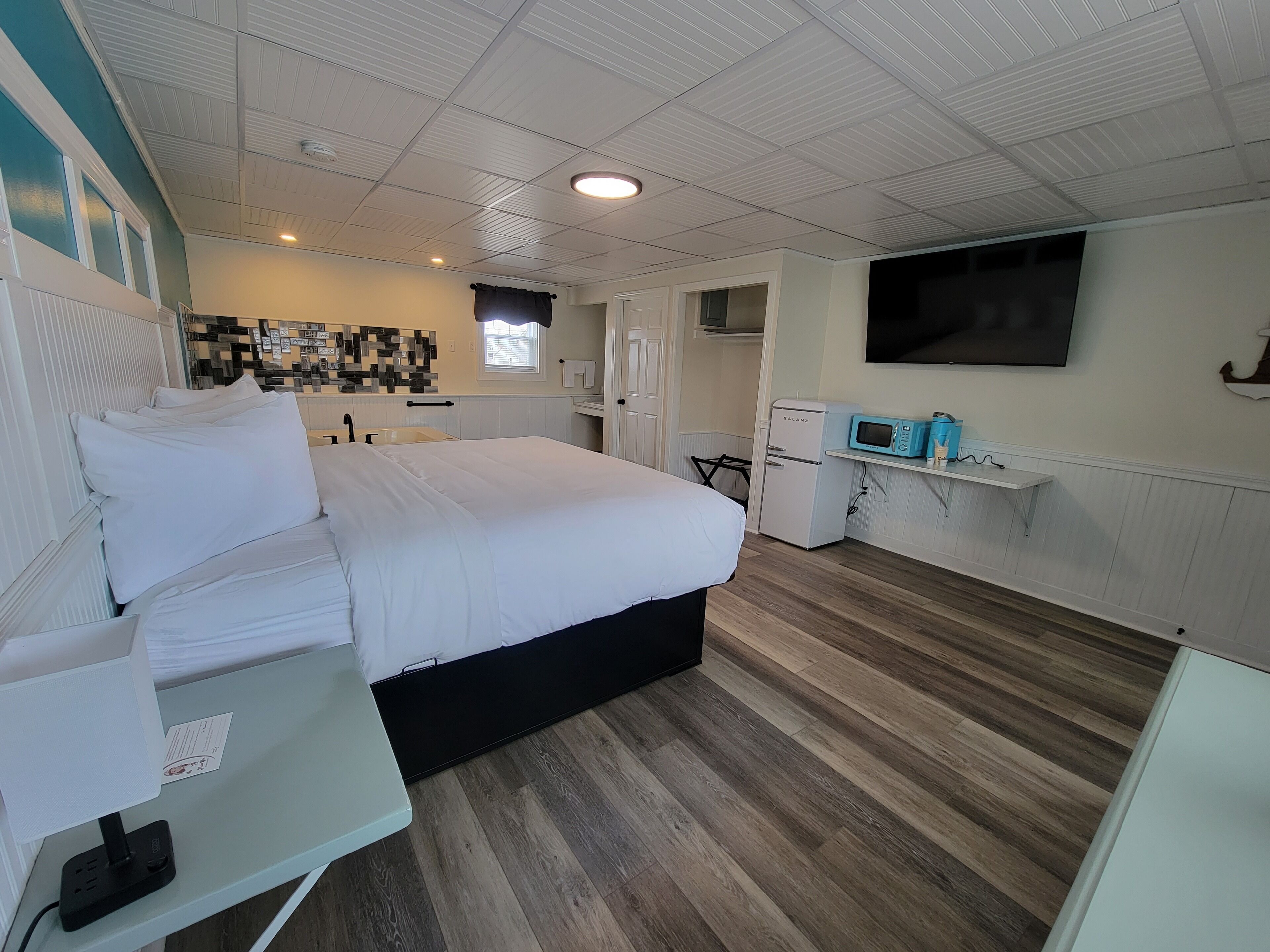 A bedroom with a bed, bedside table, and a small desk with a lamp. There is a microwave and a refrigerator in the room.