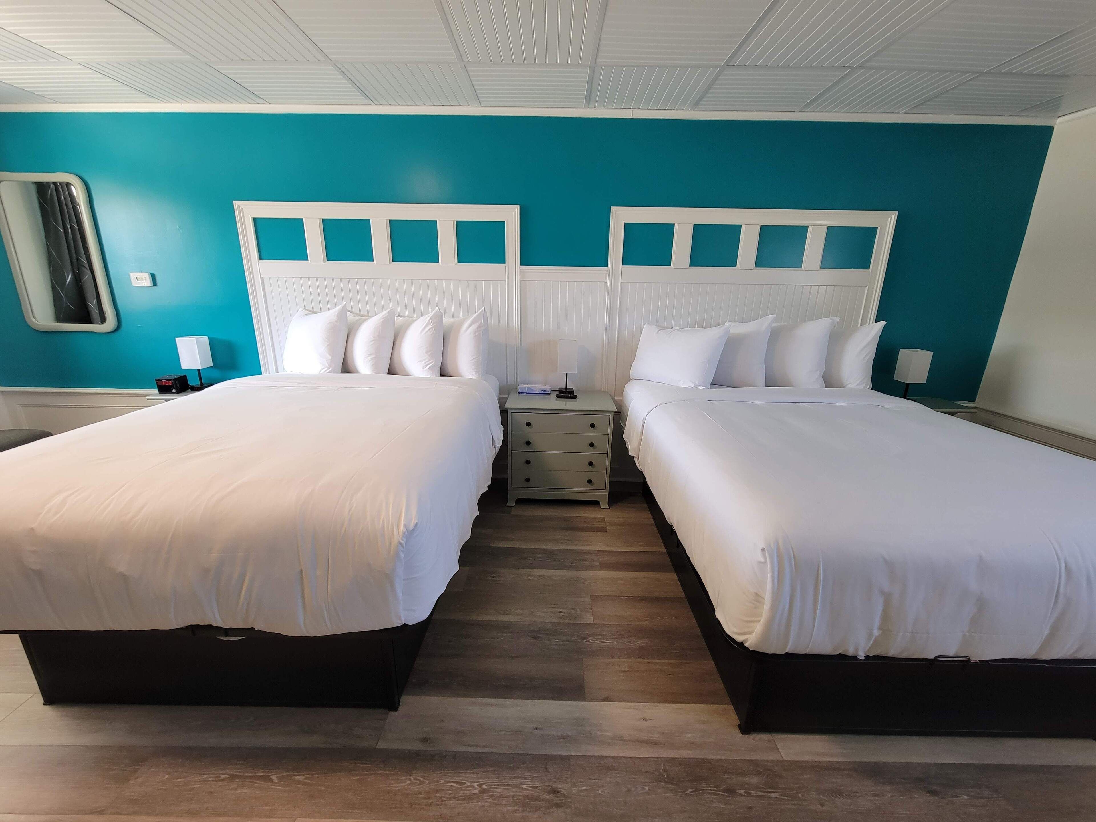 A hotel room with two beds, a nightstand, a mirror, and a teal-colored wall.