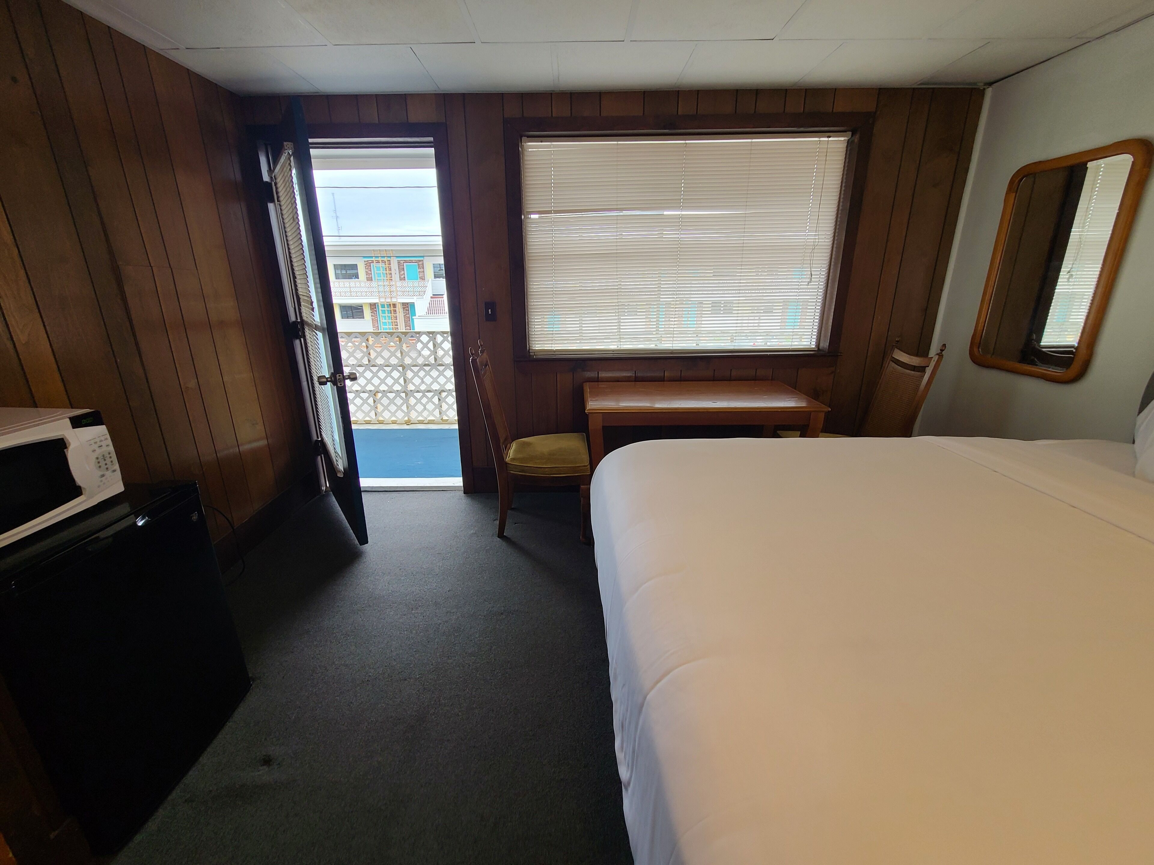 A hotel room with a bed, a desk, a chair, a microwave, and a large window with blinds.