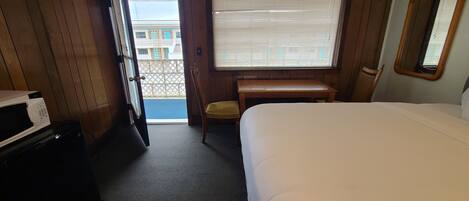 A hotel room with a bed, a desk, a chair, a microwave, and a large window with blinds.