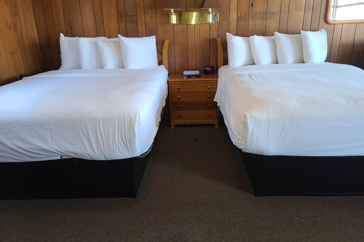 A room with two beds, a wooden floor, a nightstand with a lamp, and a window.