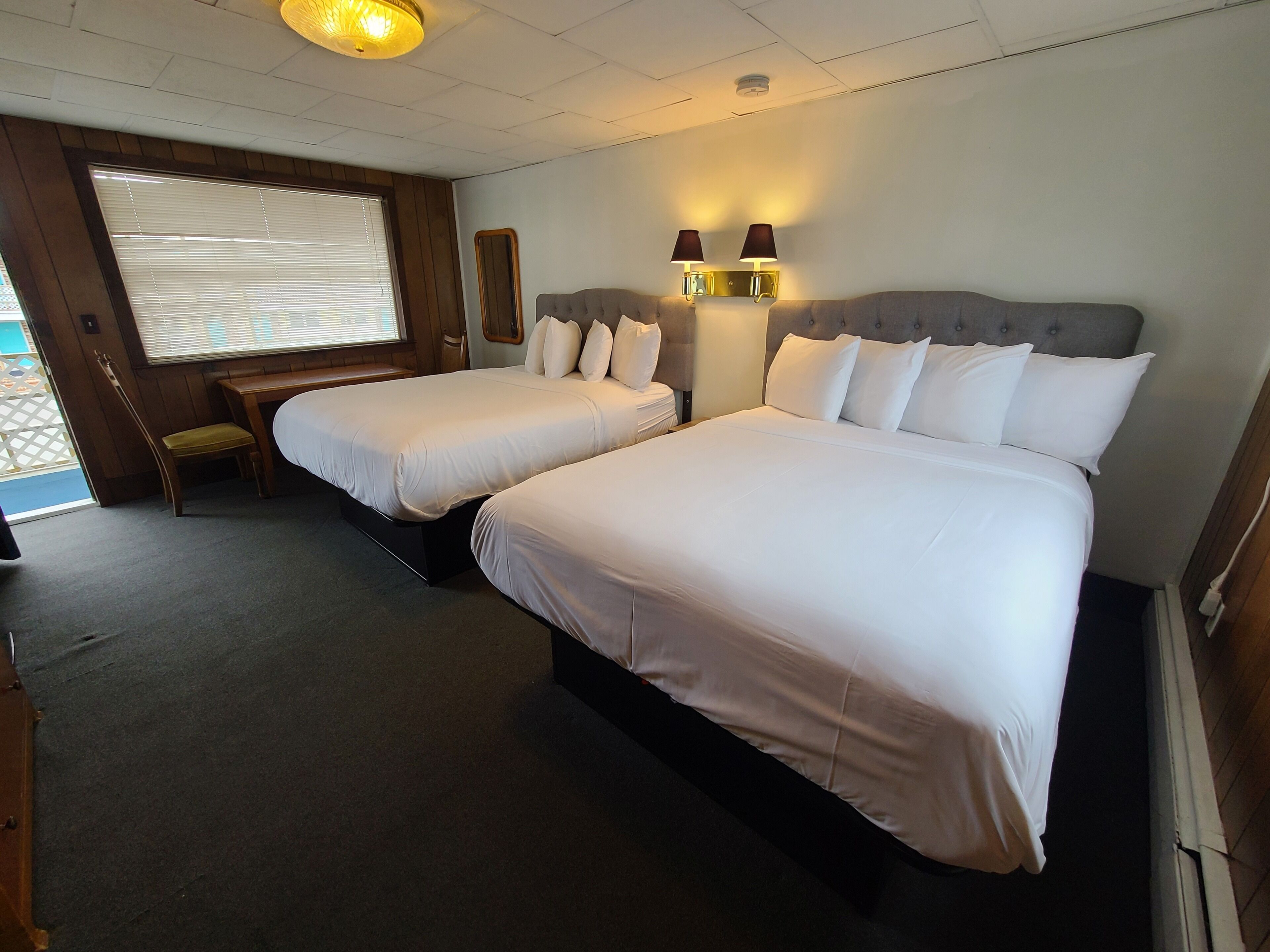 A hotel room with two beds, a wooden bench, a window with blinds, and wall-mounted lamps.