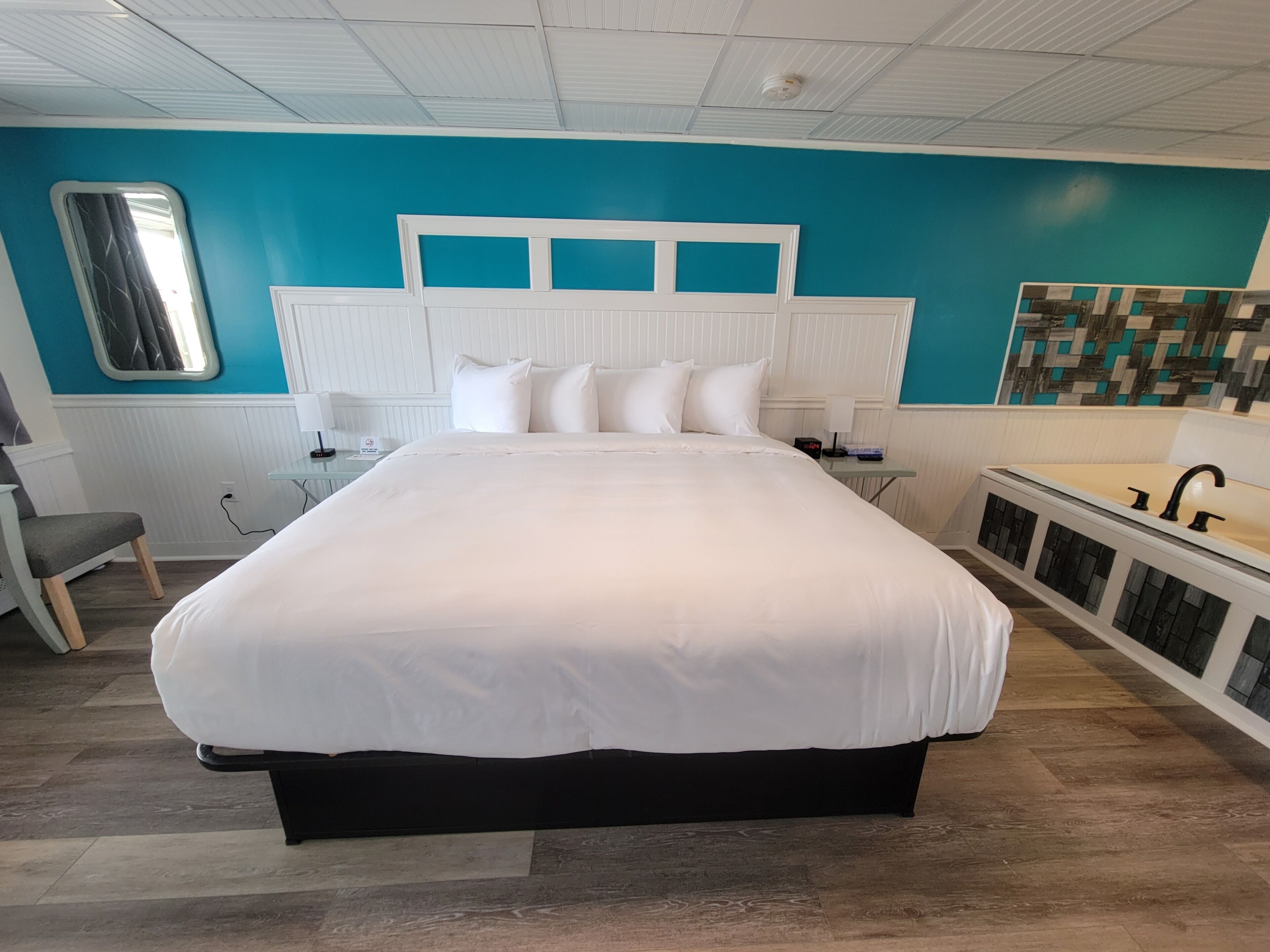 A modern bedroom with a large bed, a bedside table, a chair, a window, and a bathroom with a sink and shower.