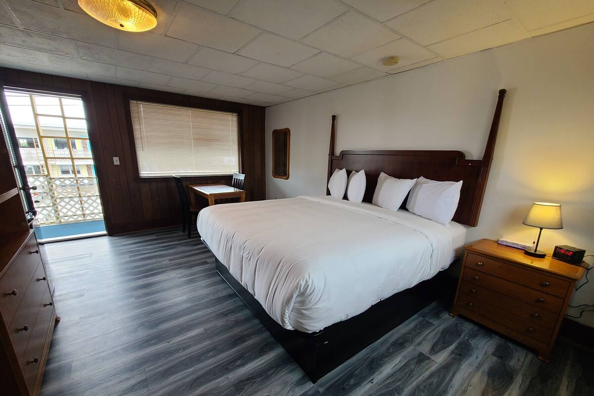 A hotel room with a large bed, a desk, and a chair. There is a window with blinds, a wooden floor, and a nightstand with a lamp.