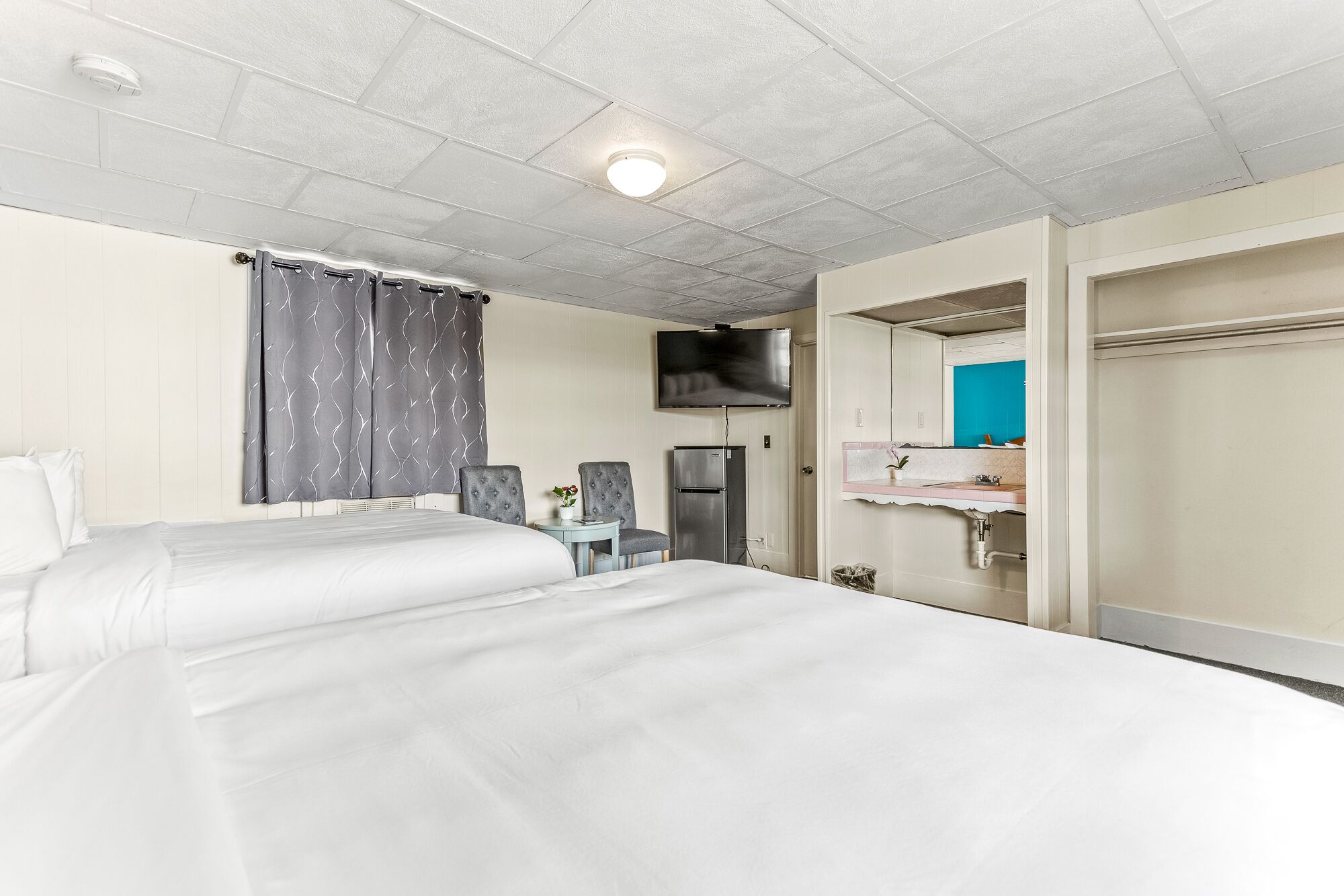 Habitación de hotel con dos camas, televisor, refrigerador, una mesita con silla y baño con lavamanos y espejo.