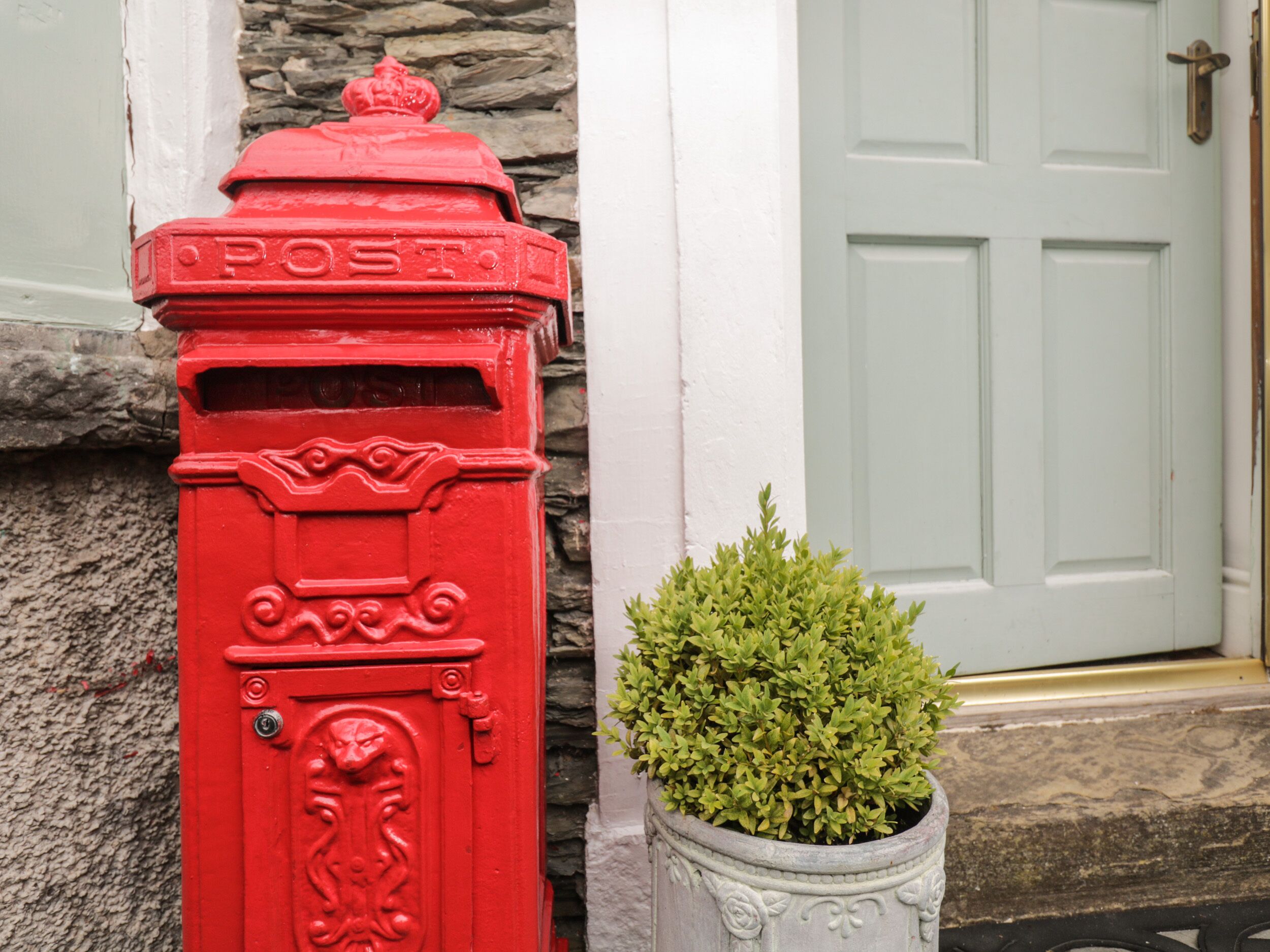 A red post box next to a white door with a stone wall and a potted plant.
