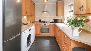 A compact kitchen with wooden cabinets, a stainless steel refrigerator, a built-in washing machine, a microwave, and a sink. - The Postmasters House (Windermere)