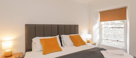 A neatly made bed with a grey headboard and two orange pillows, a bedside lamp, and a window with a view of stone walls.