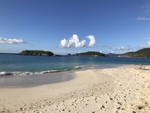 En sandstrand med fodspor, roligt vand og fjerne øer under en klar himmel.