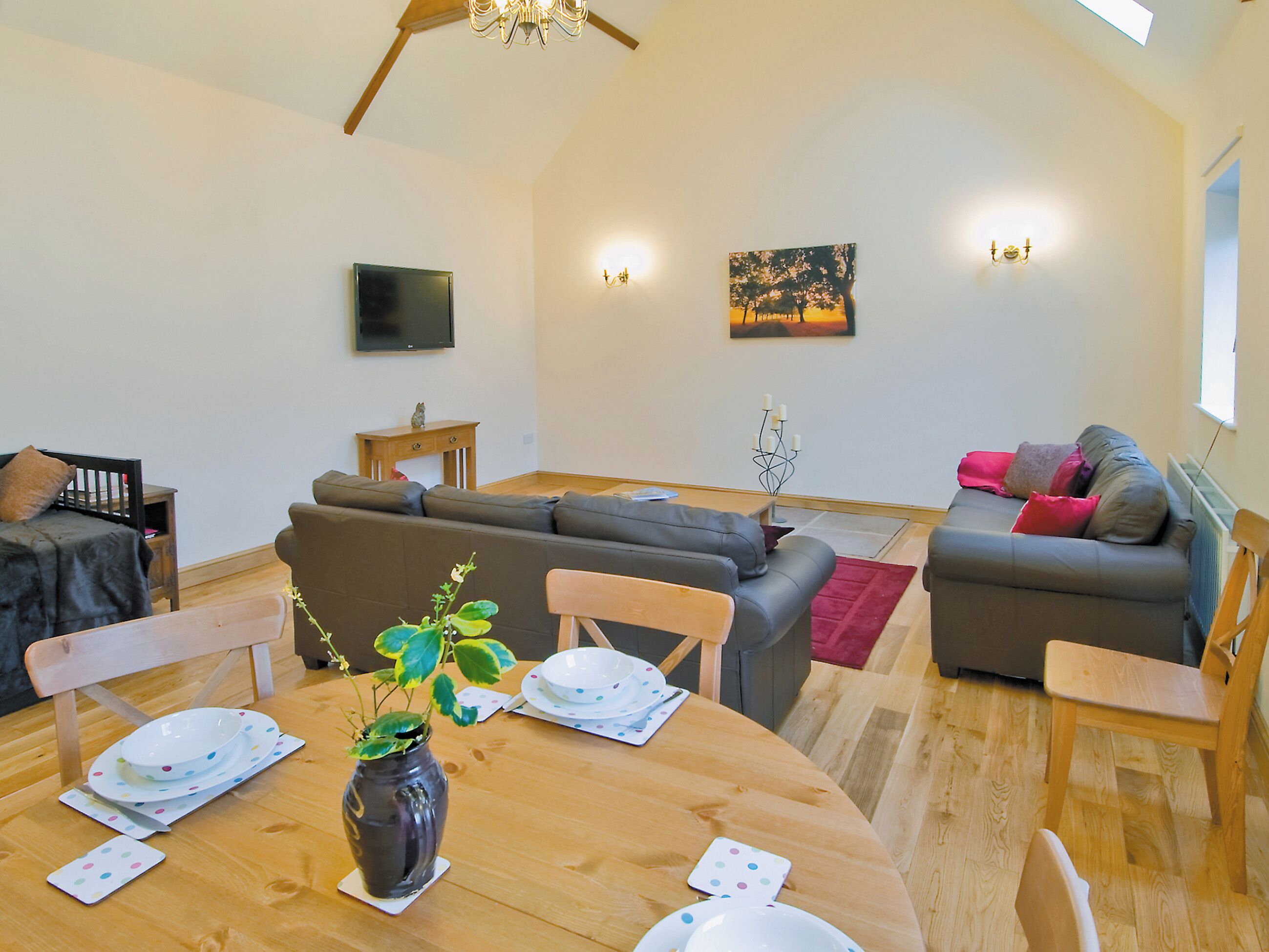 A cozy living room with a wooden dining table, grey sofas, and a TV mounted on the wall.