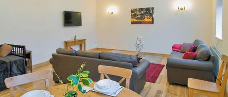 A cozy living room with a wooden dining table, grey sofas, and a TV mounted on the wall.