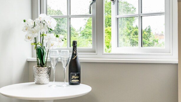 Una mesa con una botella de champán, dos copas y un florero con flores blancas, cerca de una ventana con vistas al verdor.