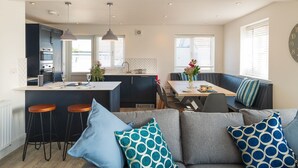 Een moderne keuken met donkere kastjes, een centraal eiland en hanglampen. Er is een eethoek met een houten tafel en blauw gestreepte kussens.