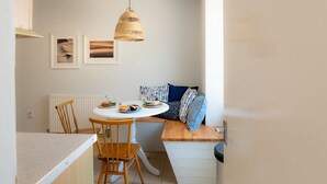 A compact kitchen with a round table, two wooden chairs, a hanging light fixture, and a radiator. - Bryn Awel Bach: 2-Bed Apartment in Central Rhosneigr | Managed by Anglesey Holiday Lettings (Rhosneigr)