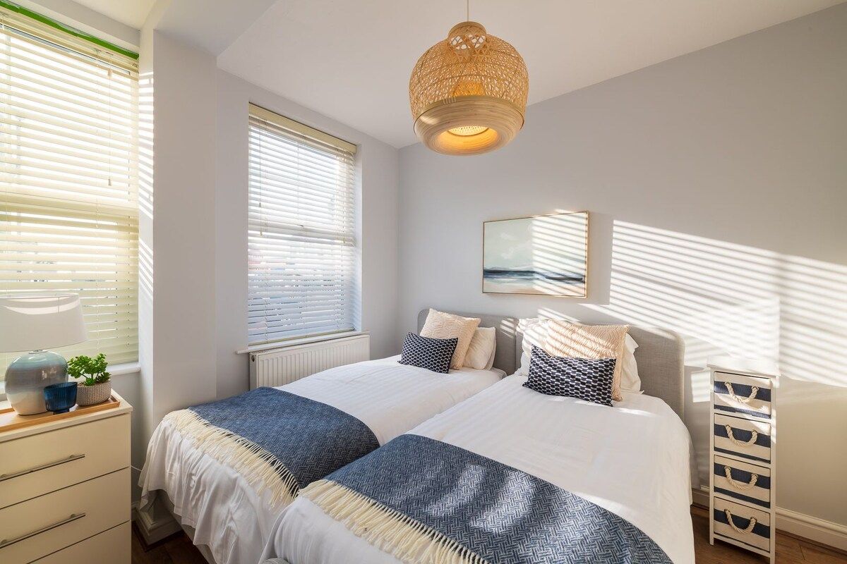 A bedroom with two beds, a bedside table, a lamp, and a window with blinds.