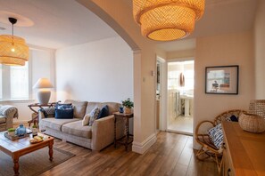 A living room with a sofa, coffee table, wicker chairs, and a dining table. There is a lamp, a framed picture on the wall, and a window with blinds. - Bryn Awel Bach: Two bedroom holiday apartment in Rhosneigr (Rhosneigr)