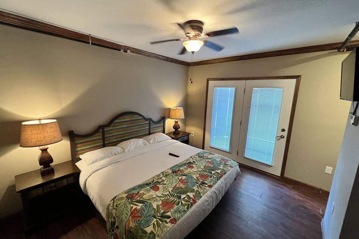 A bedroom with a bed, two bedside lamps, a ceiling fan, and a door with blinds.