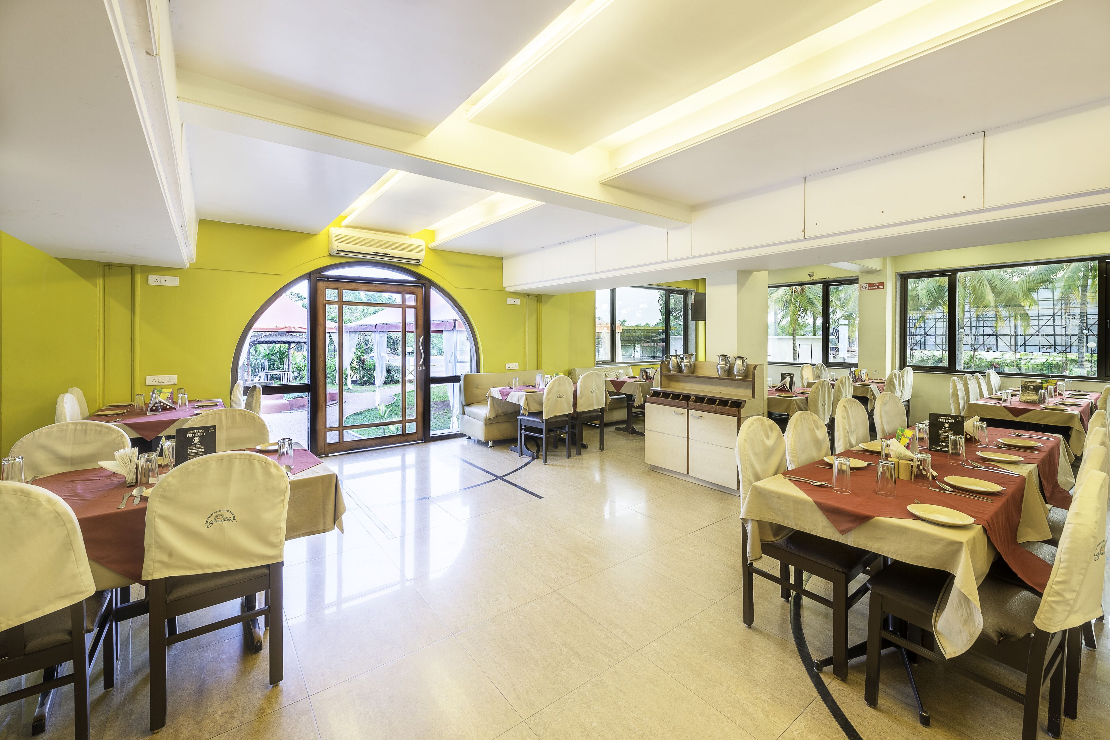 A dining area with tables set for service, a kitchen in the background, and a large window with a view of palm trees.