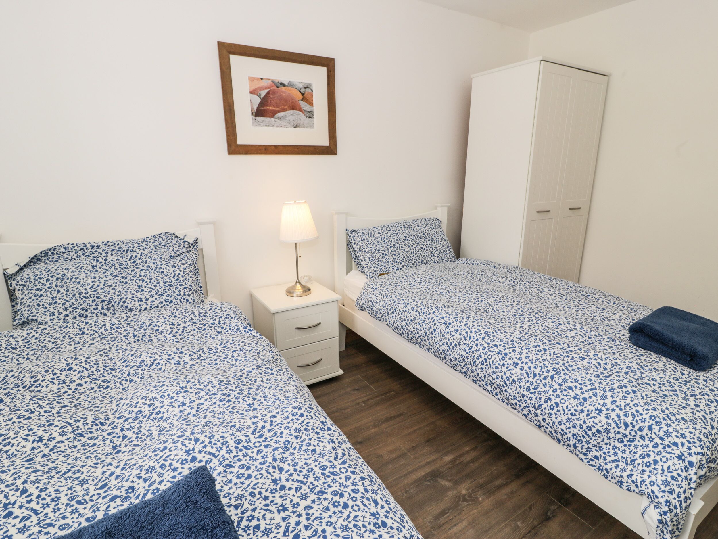 A bedroom with two beds, a bedside table with a lamp, a wardrobe, and a framed picture on the wall.