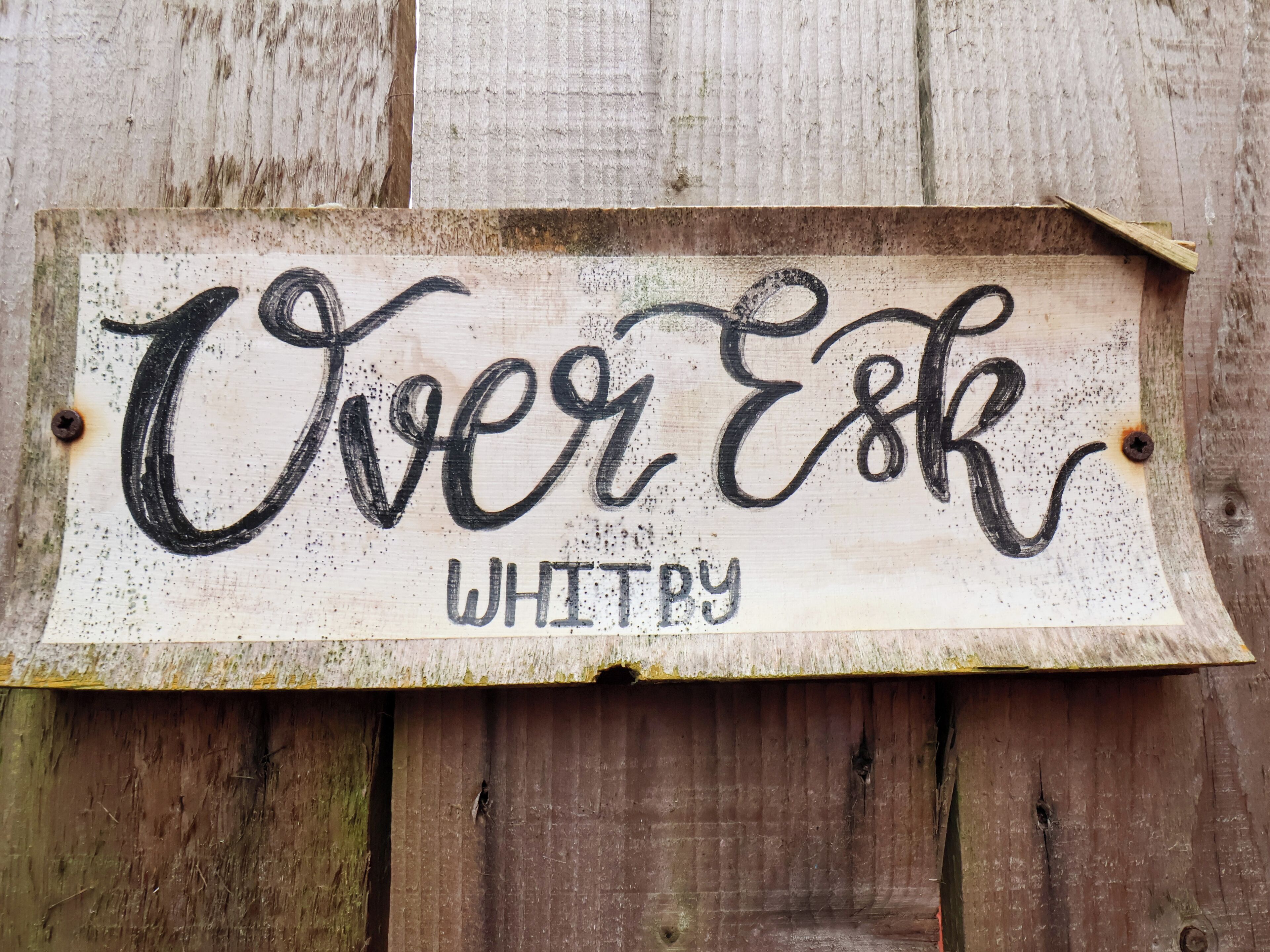Une enseigne en bois rustique sur laquelle on peut lire, dans une police d’écriture décorative, le texte « Over Esk WHITBY ».