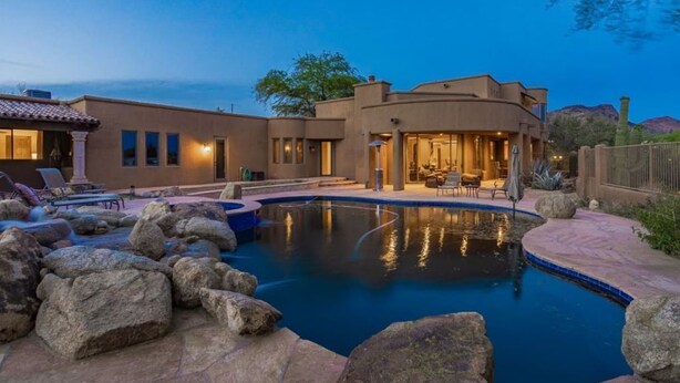 Una casa moderna con piscina rodeada de rocas, un patio con mobiliario exterior e interiores luminosos.