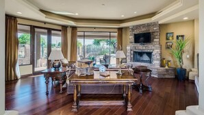 Un salón comedor amplio con chimenea, un televisor grande montado en una pared de piedra y vistas al exterior a través de grandes ventanales.