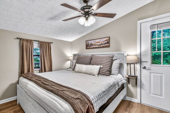 A bedroom with a bed, ceiling fan, window with curtains, bedside lamps, and a door.