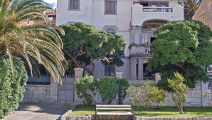 Um edifício de dois andares com varanda, rodeado por vegetação exuberante e palmeiras.