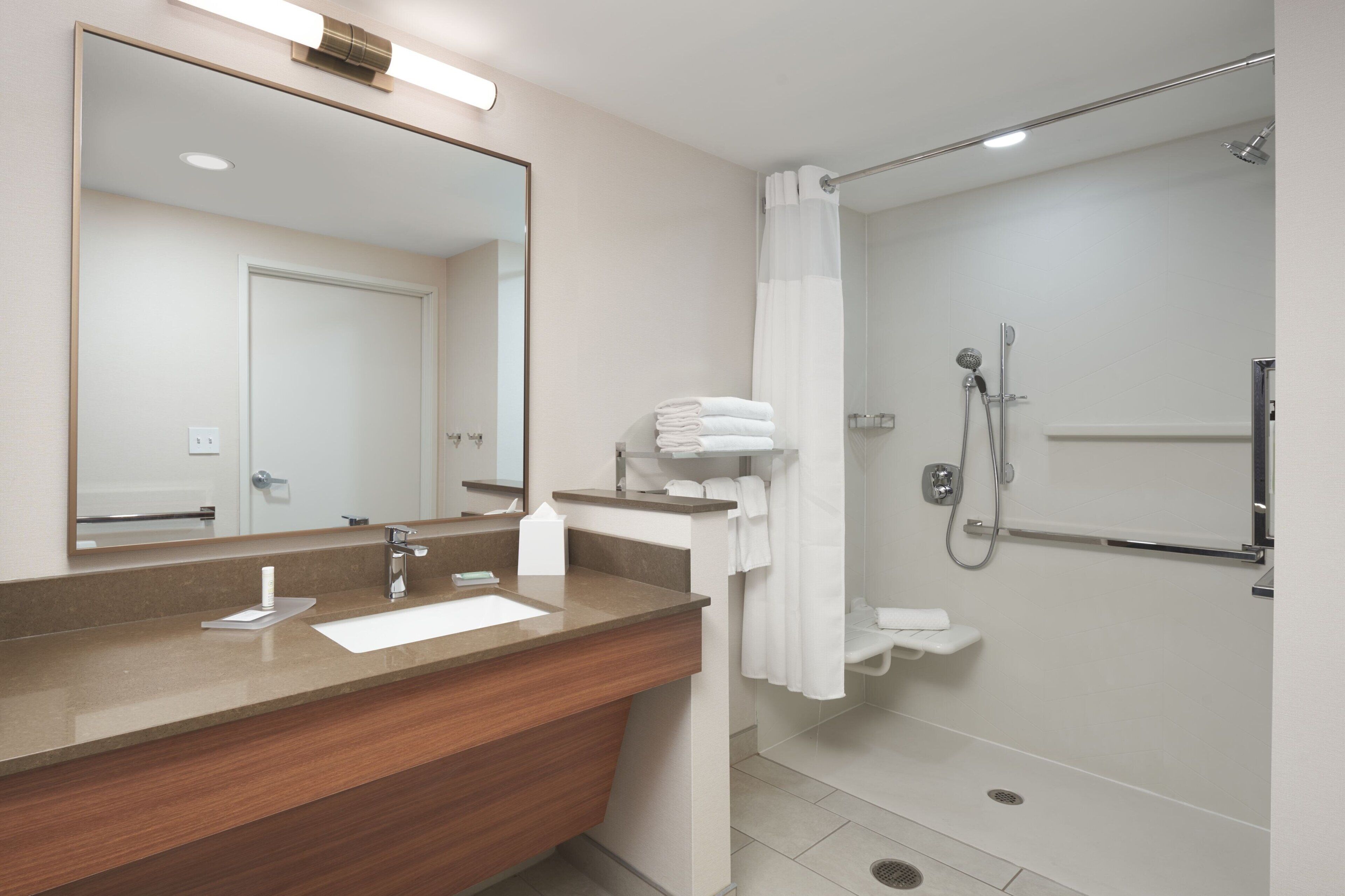 A modern bathroom with a wooden vanity, a large mirror, a shower with a handheld showerhead, and a towel rack.