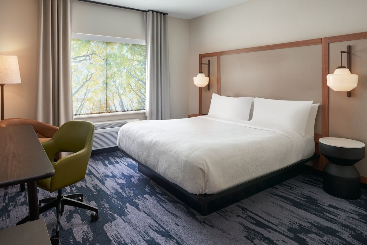 Habitación de hotel con una cama grande, un escritorio con silla, una ventana con cortinas y dos lámparas de pared.