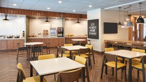 A modern restaurant with wooden flooring, yellow chairs, and tables. There is a bar area with a refrigerator and a chalkboard with the text "LESS MONDAY MORE SUMMER".