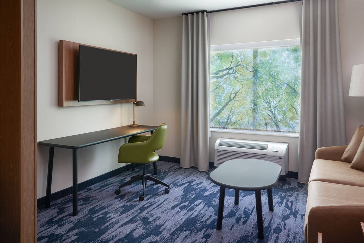 Habitación de hotel con escritorio, silla y sofá. Hay un televisor de pantalla plana en la pared, una ventana con cortinas y un equipo de aire acondicionado.