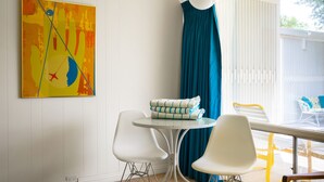A room with a white wall, a colorful abstract painting, a round table with two white chairs, a blue curtain, and a view of the outdoors.