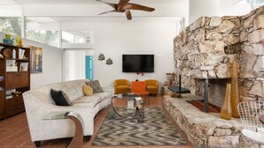 A modern living room with a stone fireplace, a white sofa, orange chairs, and a flat-screen TV mounted on the wall.