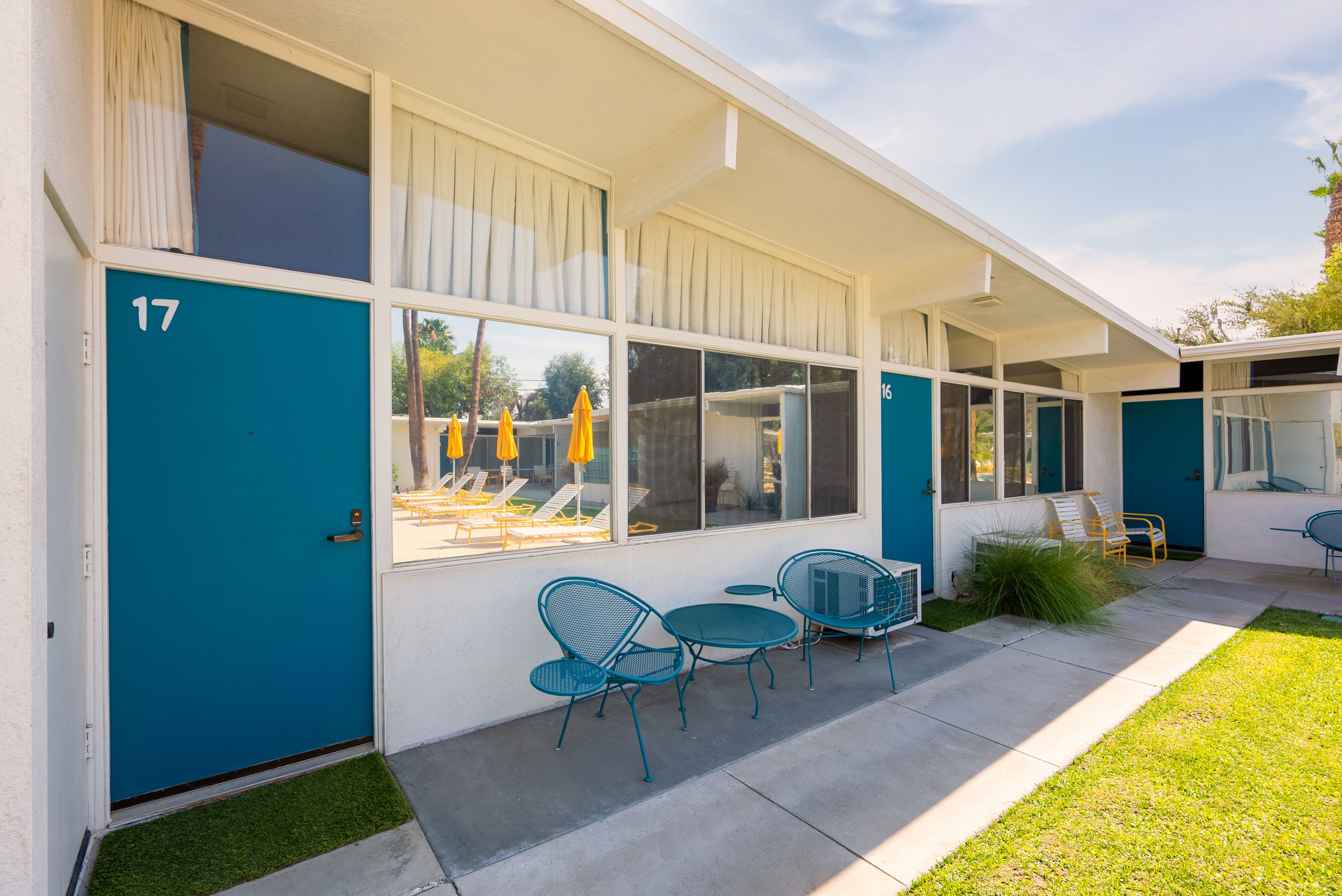 A modern building with a teal door marked '17', outdoor seating with yellow umbrellas, and a clear view of the pool area.