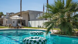A swimming pool with two checkered inflatable rings, lounge chairs, and a palm tree.
