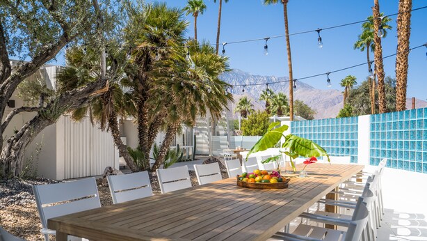 An outdoor dining area with a wooden table and white chairs, surrounded by palm trees and a mountain view.