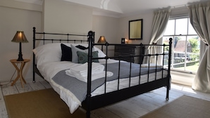 A neatly made bed with a black frame, a bedside lamp, and a dresser in a room with a large window and a view of the outside. - Matchbox is a delightful dog-friendly cottage in Winchelsea, East Sussex (Winchelsea)