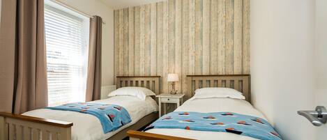 A bedroom with two single beds, a nightstand with a lamp, a window with curtains, and a striped wallpaper on the wall.