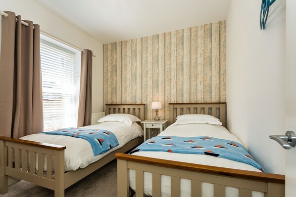 A bedroom with two single beds, a nightstand with a lamp, a window with curtains, and a striped wallpaper on the wall.
