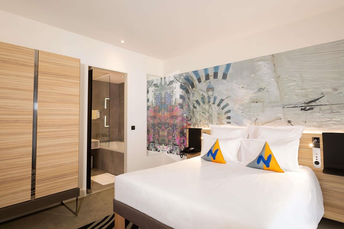 A hotel room with a bed, a bathroom, and a large abstract painting on the wall.