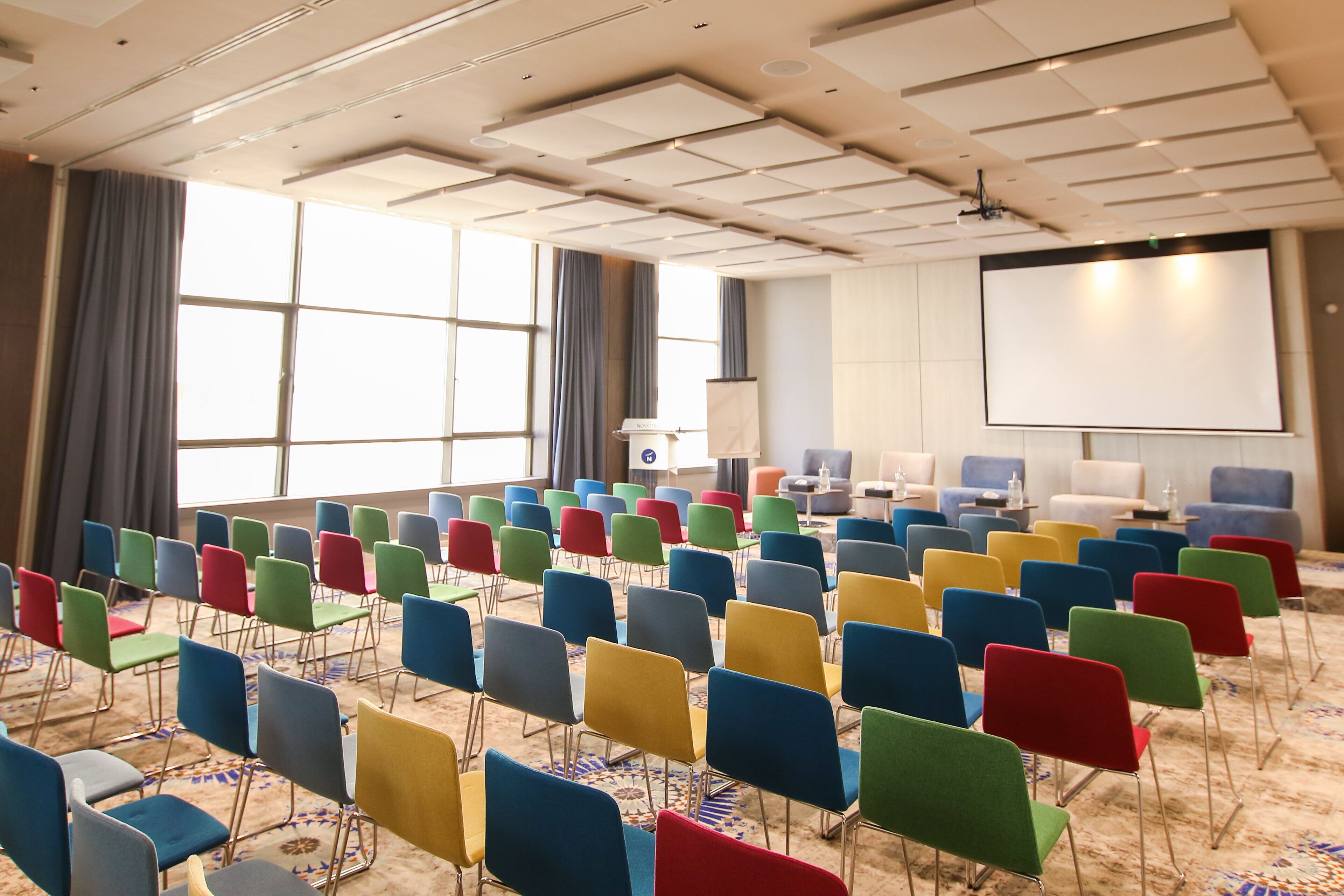 Un salón de conferencias con filas de sillas de colores, una pantalla grande y un proyector.