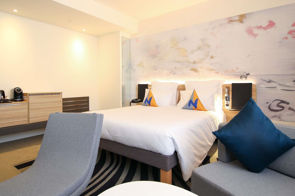 A modern hotel room with a large bed, a desk with a coffee maker, a seating area with a sofa and armchairs, and a wall mural.
