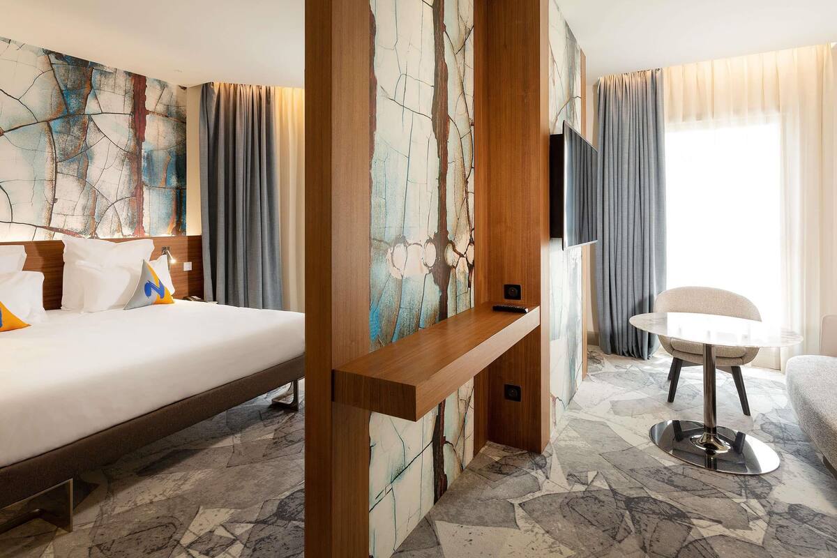 A hotel room with a bed, a desk, a chair, a TV, and a large window with curtains.
