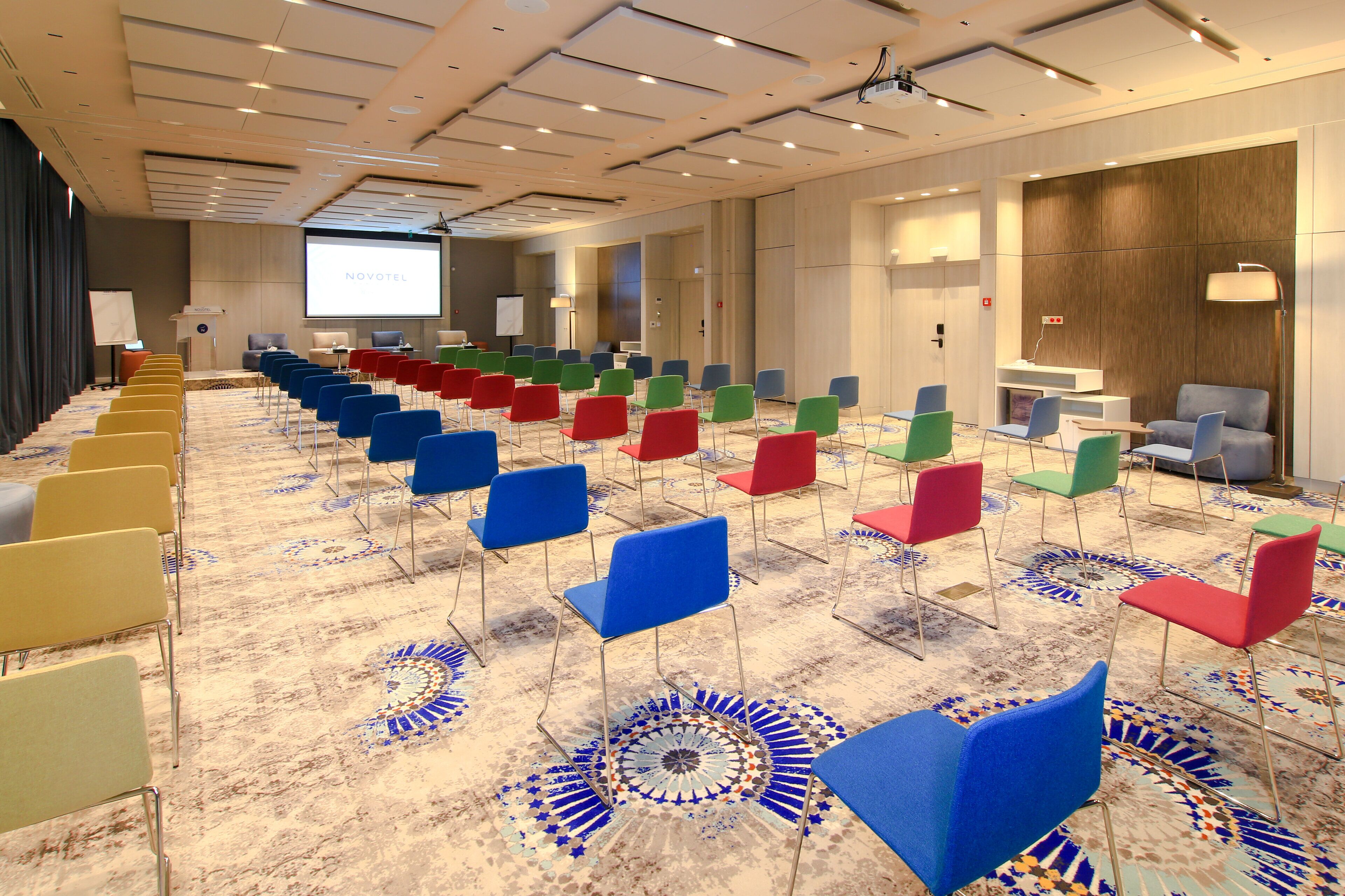 Une salle de conférence avec des rangées de chaises, un écran de projection et une moquette ornée d’un motif géométrique.