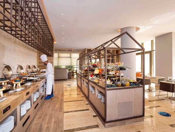 Un restaurante de hotel moderno con un chef preparando alimentos, un bufé con diversos platos y un área de estar con mesas y sillas.