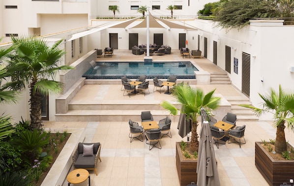 A rooftop pool area with multiple seating arrangements, including sofas and tables, surrounded by palm trees and plants. - Novotel Tunis Lac (Tunis)
