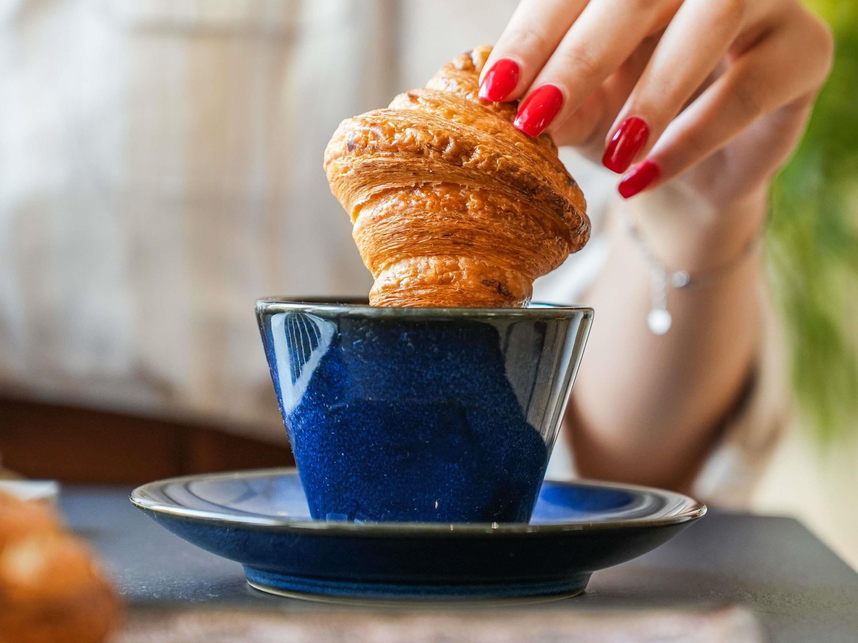Eine Hand hält einen Croissant über einer blauen Keramiktasse.